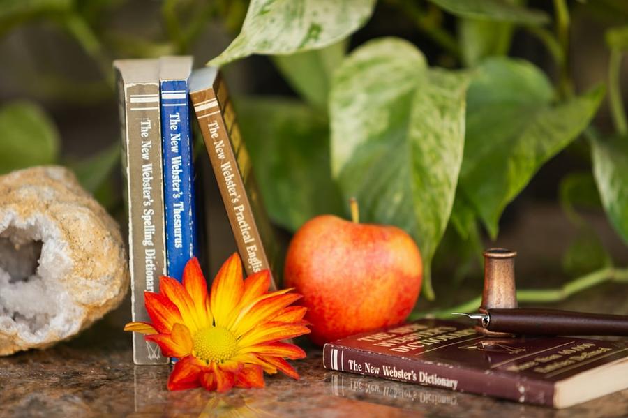A well-worn thesaurus and dictionaries stand beside an inkwell, apple, and flower.