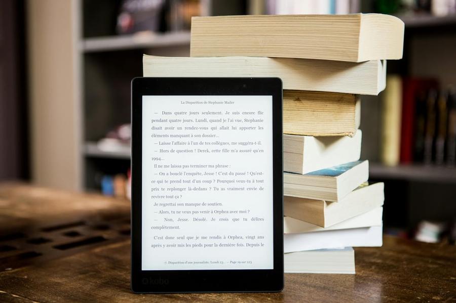 An e-reader with an e-book open on the screen leans against a stack of physical books.