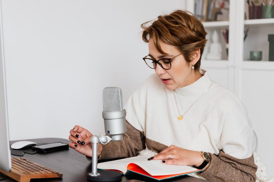 A person sits at a table reading a book aloud into a microphone.