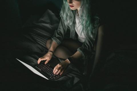 A writer with teal hair types on a laptop in a dark room.