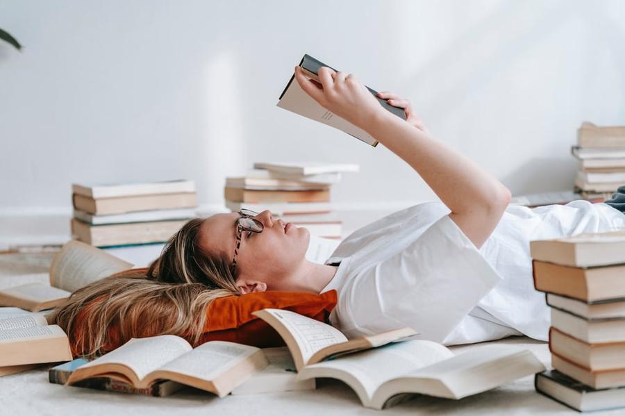 A person lies on their back surrounded by books and reading the first line of a book.
