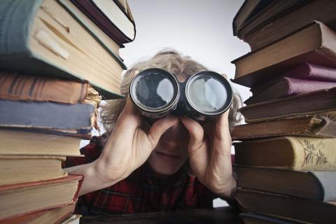 A person looks through two stacks of books with a pair of binoculars.