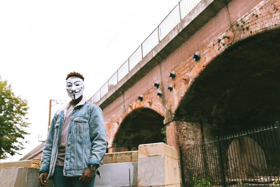 A person in a jean jacket and a Guy Fawkes mask standing in front of a bridge.