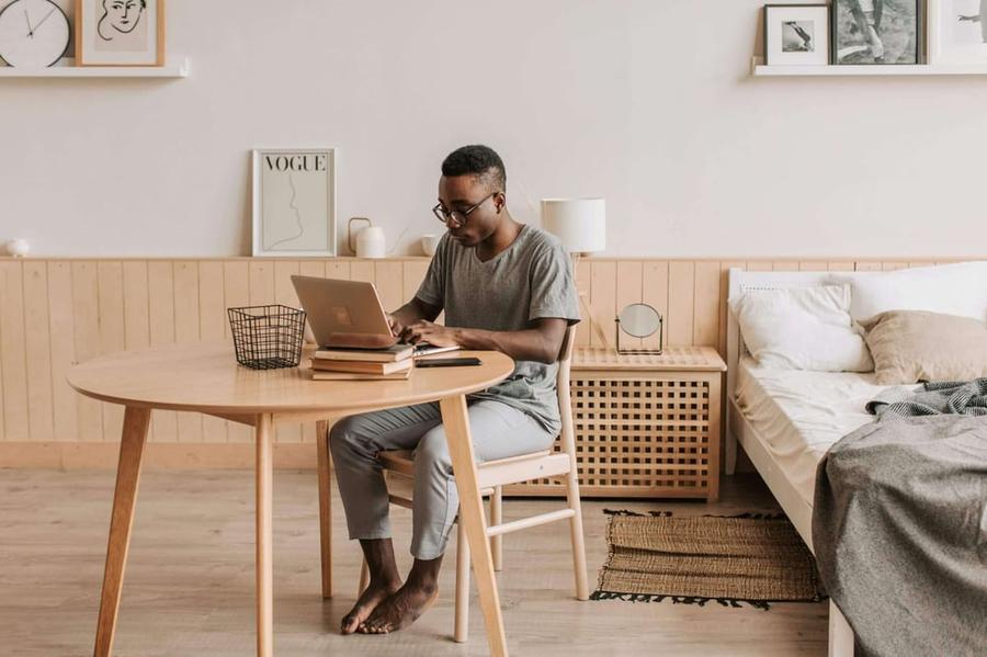 Writer typing on a laptop next to a stack of books on a round table in a bedroom.