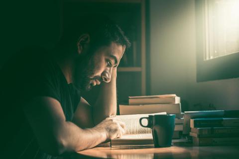 A person sits at a desk in a dark room surrounded by books and reads by the light streaming in through the window.