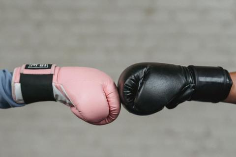 Two hands butt up against one another, one in a black boxing glove and one in a pink boxing glove.