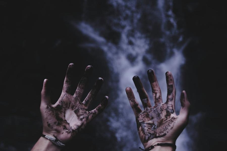 Hands coated in a black substance reach out against a dark and smokey background.