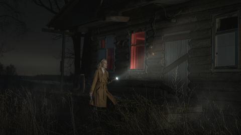 Woman in a brown trench coat with a flashlight standing in front of an abandoned house at night.