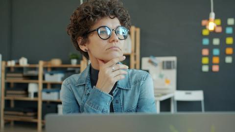 A person with short curly hair and glasses sits in front of a computer, looking off into the distance as if in thought.