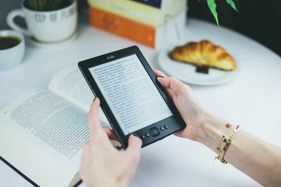A pair of hands holds an e-reader displaying an e-book at a desk.