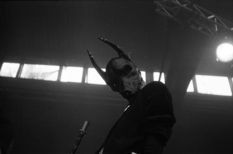 A person wearing an ambiguous animal mask with horns stares down at the viewer.