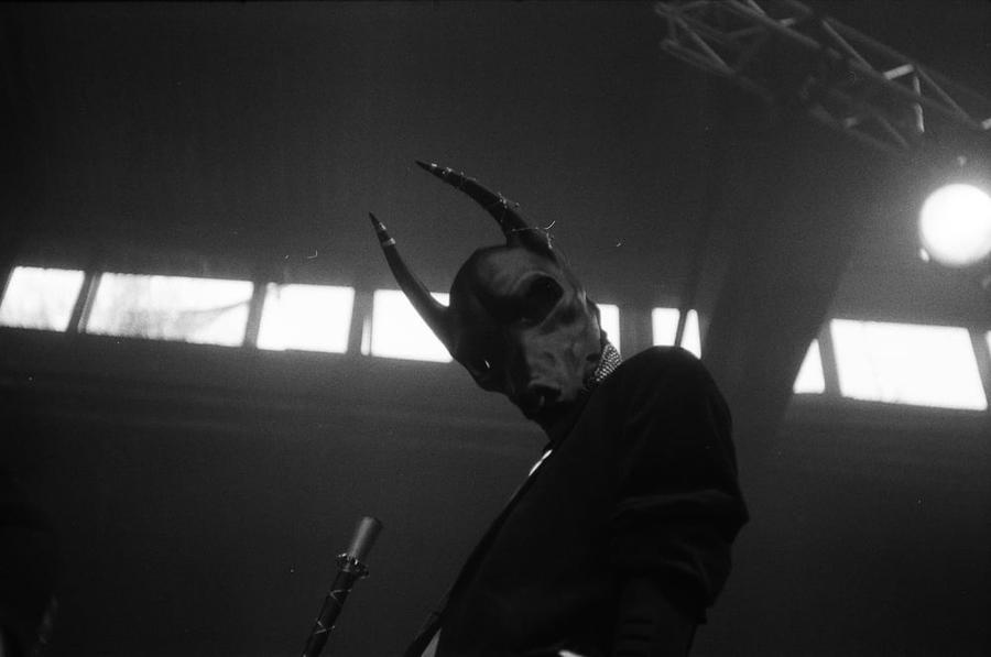 A person wearing an ambiguous animal mask with horns stares down at the viewer.