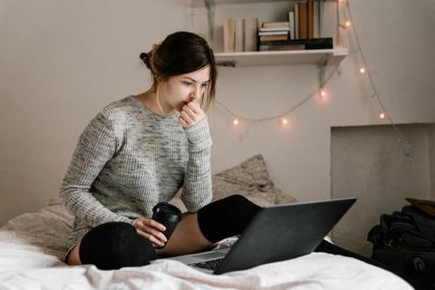 A person sits on a bed, staring at a laptop screen and thinking.