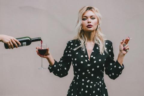 A person holds a cigarette in one hand and a wine glass in the other. A hand reaches out to pour wine in the wine glass.