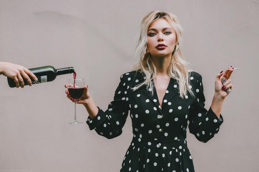 A person holds a cigarette in one hand and a wine glass in the other. A hand reaches out to pour wine in the wine glass.