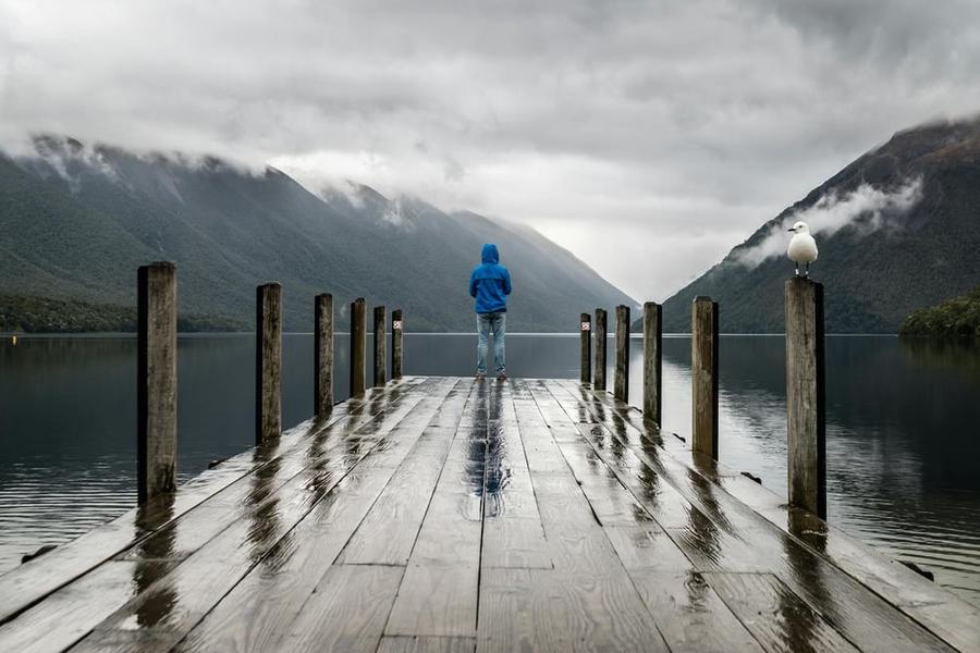 A person standing at the end of a dock, looking out at mountains.