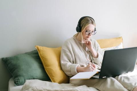 Woman sitting on a couch, headphones on, with a laptop and notebook on her lap.