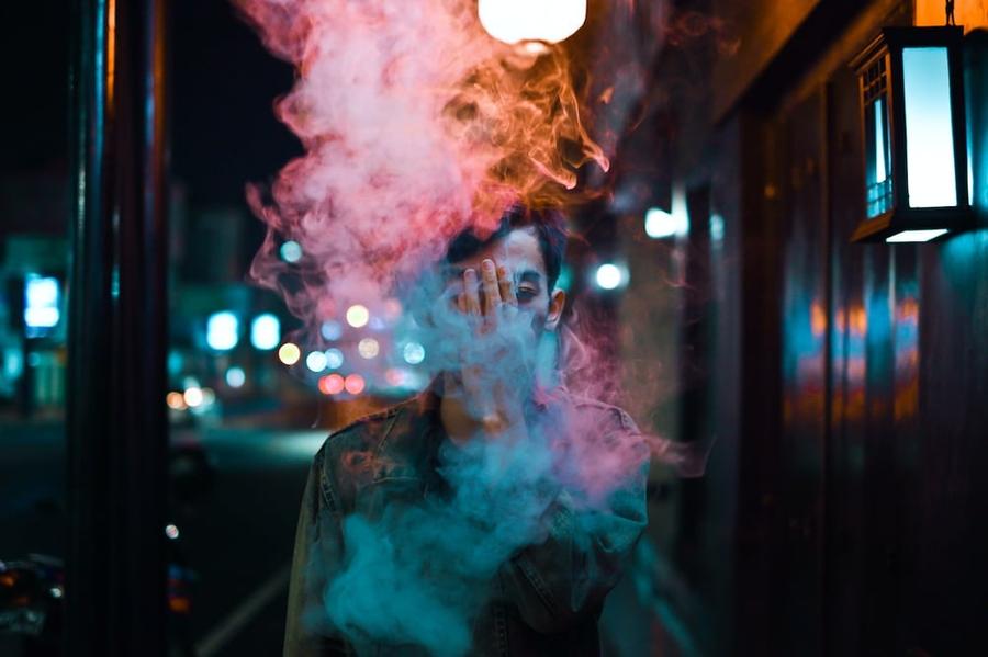 Man in a leather jacket on a city street with multi-colored smoke in front of his face.