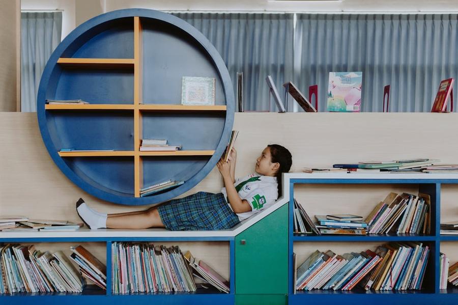 A child lies across a bookshelf, reading middle grade fiction. 