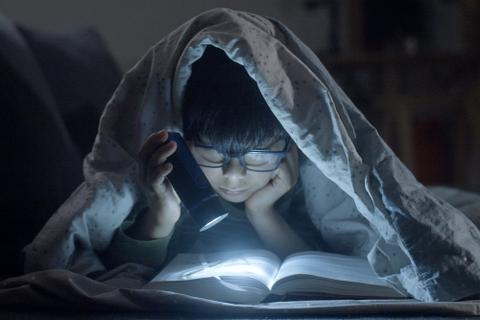 A kid wearing glasses lies on their stomach under a comforter, reading middle grade fiction by flashlight.
