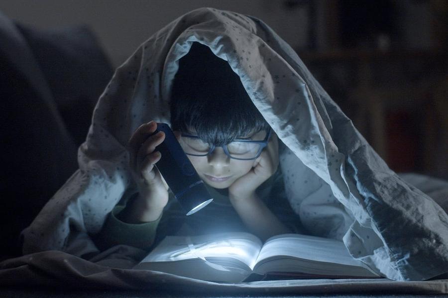 A kid wearing glasses lies on their stomach under a comforter, reading middle grade fiction by flashlight.