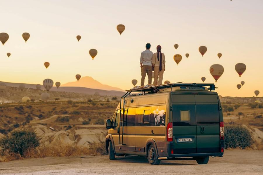 Two people stand on the roof of a vintage van in the desert, watching dozens of hot air balloons rise into the sky.