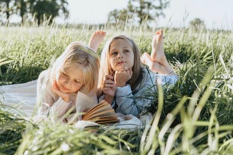 Two young girls lying on a blanket in a field of tall grass. One is reading a book while the other is staring up to the sky.