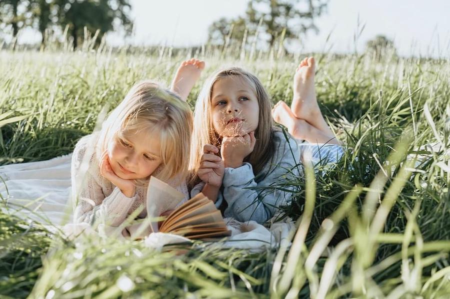 Two young girls lying on a blanket in a field of tall grass. One is reading a book while the other is staring up to the sky.
