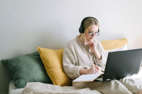 A person sits up in bed wearing headphones and writing in a notebook on top of a laptop.