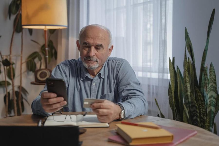 An older man holding his phone and credit card while sitting at the table.