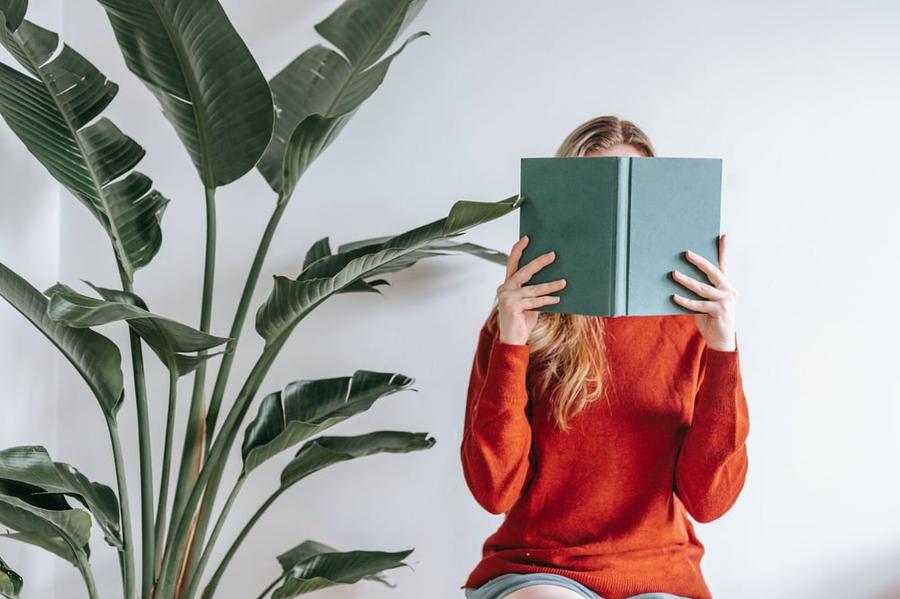 A person sitting beside tall houseplants holds an open book in front of their face.