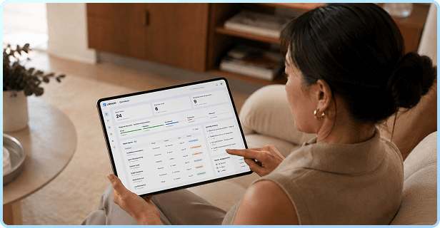 A woman sits on a couch using a tablet displaying a dashboard with charts and data in a cozy living room setting.