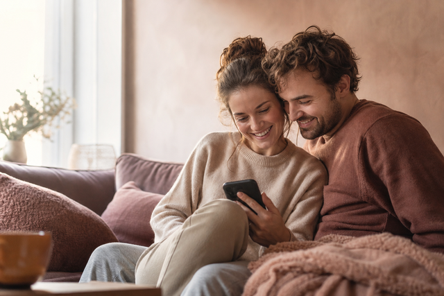 Twee partners zitten warm tegen elkaar aan op de bank en kijken samen glimlachend naar een telefoon