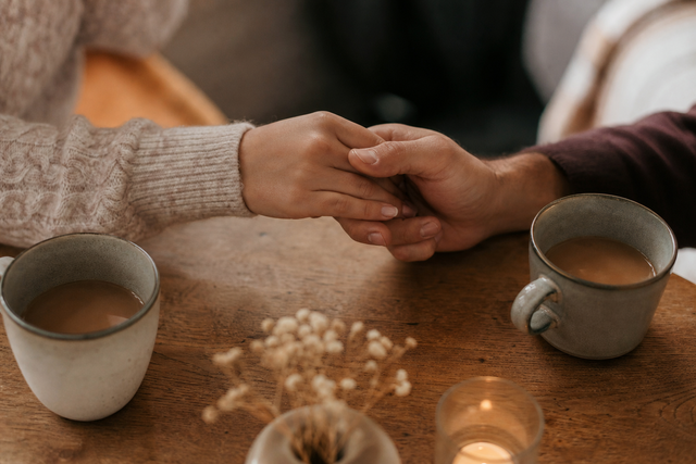 Close-up van twee handen die elkaar zachtjes vasthouden boven een kop koffie op een houten tafel
