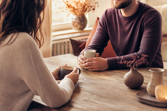 Twee partners zitten warm tegenover elkaar aan een tafel in gesprek, met kopjes koffie tussen hen in