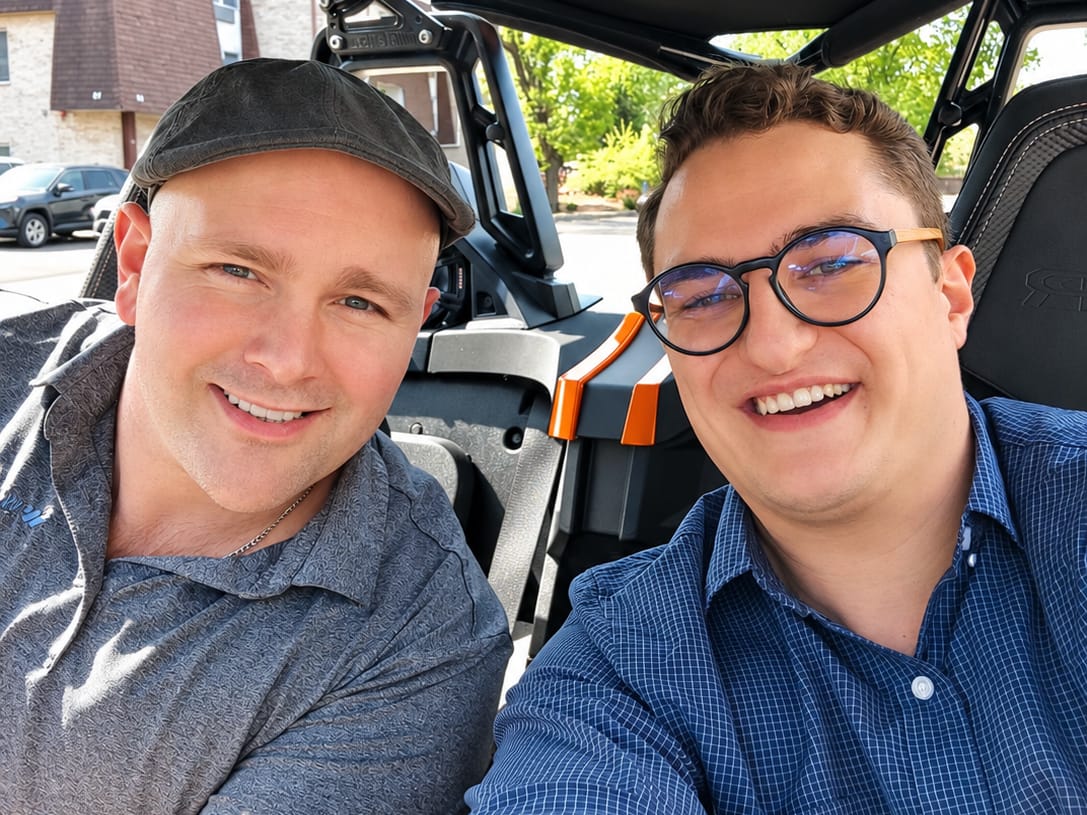 Two people smiling in a selfie from the front seats of an open off-road vehicle on a sunny day