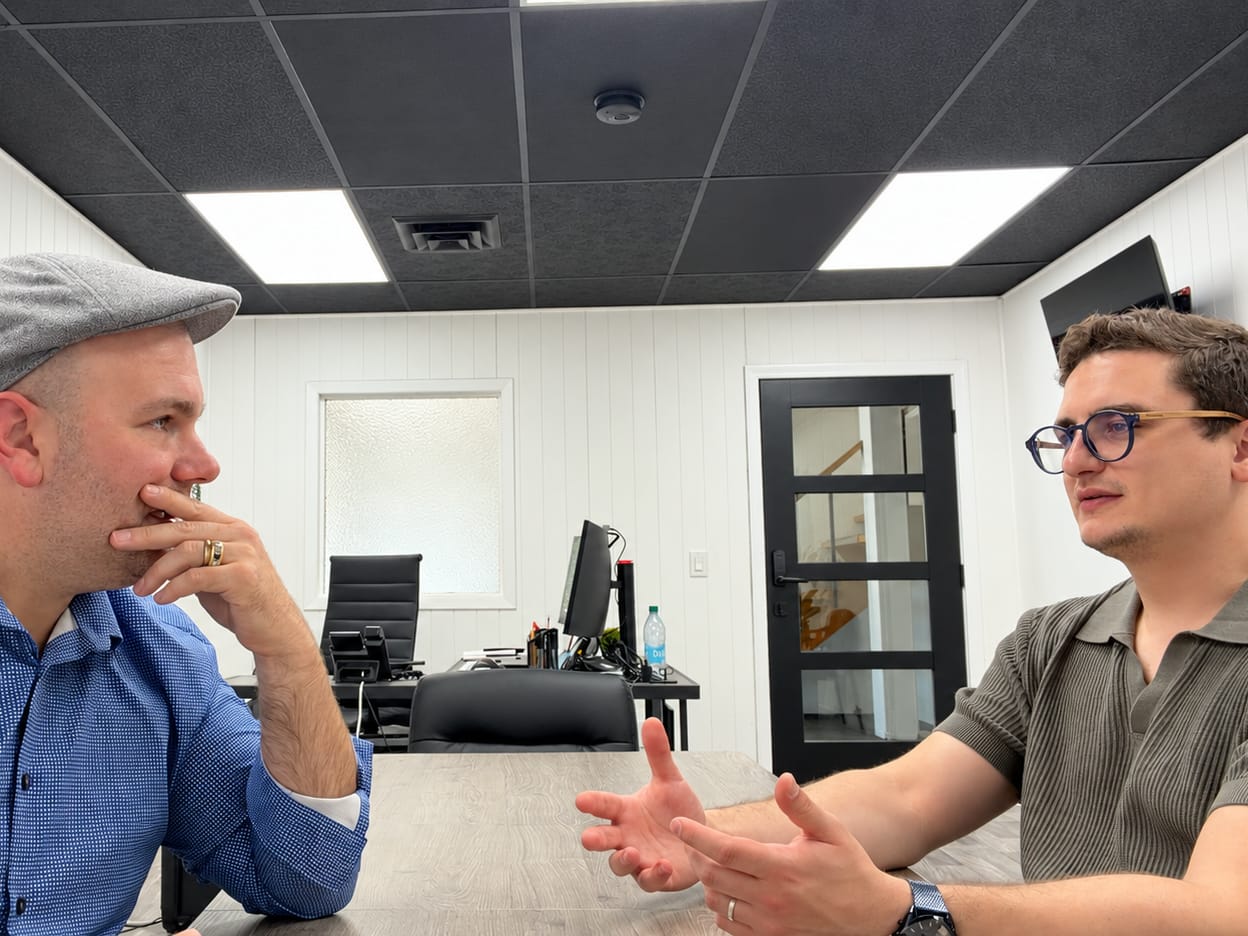 Two people in conversation across an office table, one listening with chin in hand, the other explaining with open hands