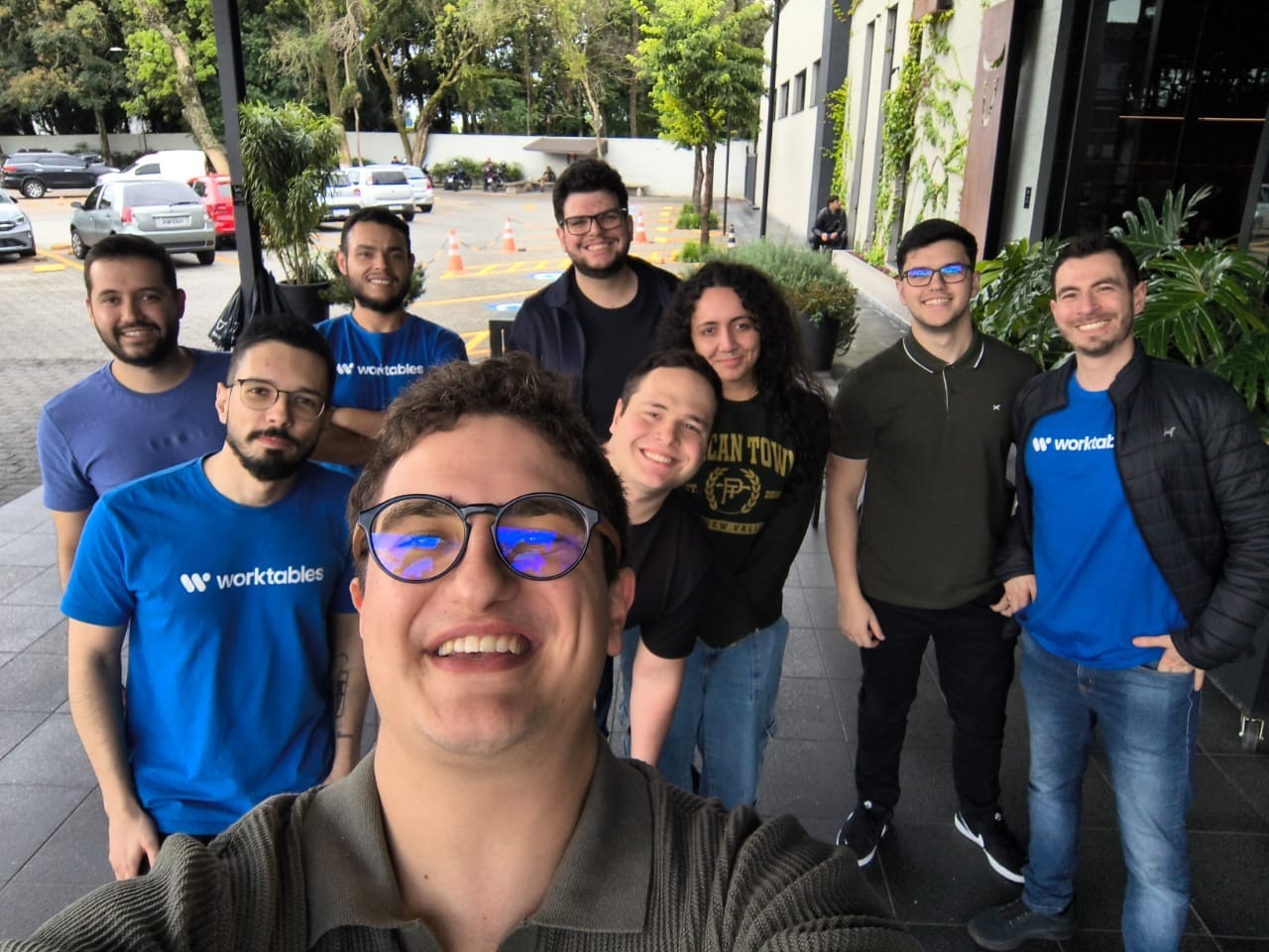 A group selfie outside an office building, several of the group in blue Worktables T-shirts