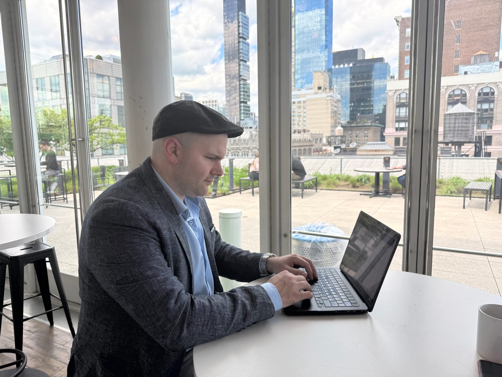 Michael at work on a laptop at a table by a wall of windows, a rooftop terrace and city towers behind him