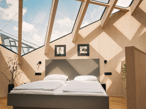 Lichtdurchflutetes Schlafzimmer im IKUNA Sternenhimmelchalet mit gemütlichem Doppelbett, heller Holzverkleidung und großzügigen Dachfenstern mit Blick in den Himmel.