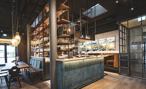Moderne Bar mit Fliesenverkleidung präsentiert Gläser und Flaschen, Holzmöbel und Beleuchtung akzentuieren stilvolle, einladende Atmosphäre.