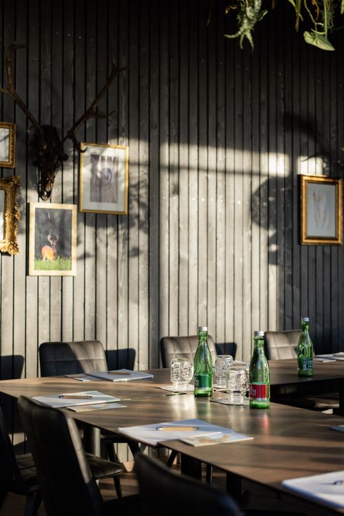 Mehrere Flaschen Wasser und Notizbücher stehen auf einem Konferenztisch in einem Raum mit Holzwand und Bilderrahmen.