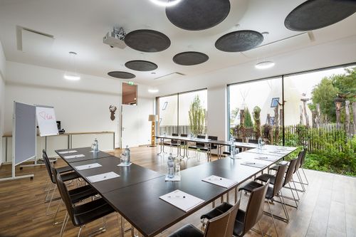 Konferenztisch mit Stühlen steht bereit. Wasserflaschen und Notizpapier sind darauf platziert. Der Raum hat große Fenster mit Blick auf einen Garten.