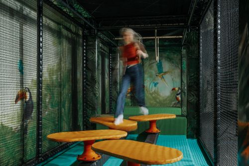 Eine Frau, dessen Bewegung durch die Kamera verwischt wirkt, balanciert über orangefarbene Trittplattformen im Netzen gesicherten Indoor-Abenteuerparcours IKUNA Ninja-Dschungel und sorgt so für eine lebhafte, verspielte Atmosphäre.
