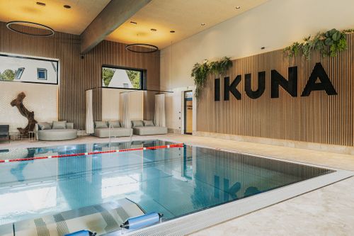Indoorpool der IKUNA Spa- & Wellnesswelt in Natternbach mit gepolsterten Daybeds, hellen Vorhängen und großen Fenstern. Pflanzen und der IKUNA-Schriftzug schmücken die holzverkleidete Wand.