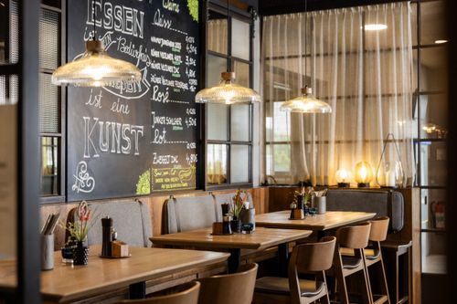 Tische und Stühle stehen in einem gemütlichen Restaurant. An der Wand eine beschriftete Tafel mit Menü. Hängelampen beleuchten den Raum.