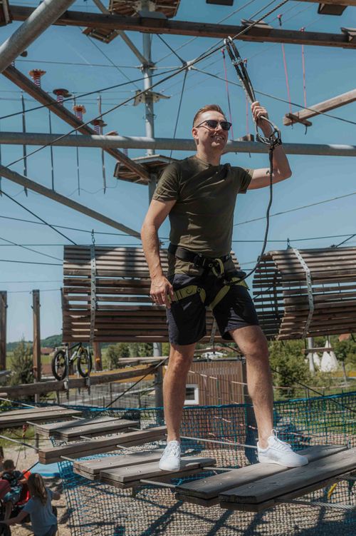 Ein Mann mit Sonnenbrille und Sicherheitsgurt bewältigt an einem sonnigen Tag im IKUNA Hochseilgarten und hält dabei ein Seil über den hängenden Plattformen fest.
