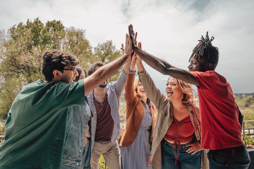Eine bunte Gruppe von Freunden, die lächelnd und jubelnd mit erhobenen Händen unter einem strahlend bewölkten Himmel im Freien ein High-Five austauschen.