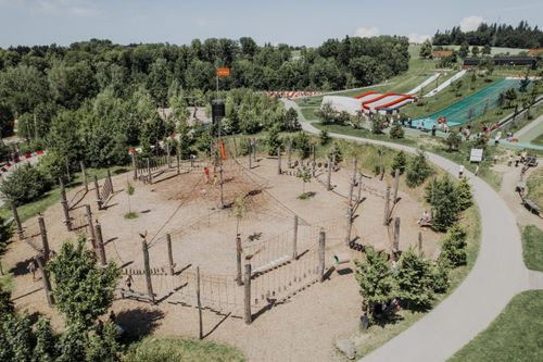 Kletterpark mit Seilen und Plattformen, umgeben von Bäumen. Besucher nutzen die Kletterelemente. Im Hintergrund eine Rutsche und Spazierwege.