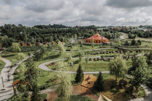 Ein Park mit Wegen, Bäumen und einem orange-roten Zelt. Dinosauriermodelle stehen verteilt. Wolkiger Himmel im Hintergrund.